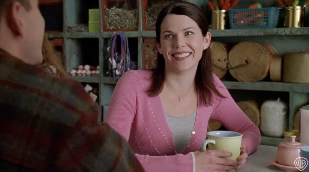lorelai gilmore of gilmore girls pictured in a pink sweater cradling a cup of coffee